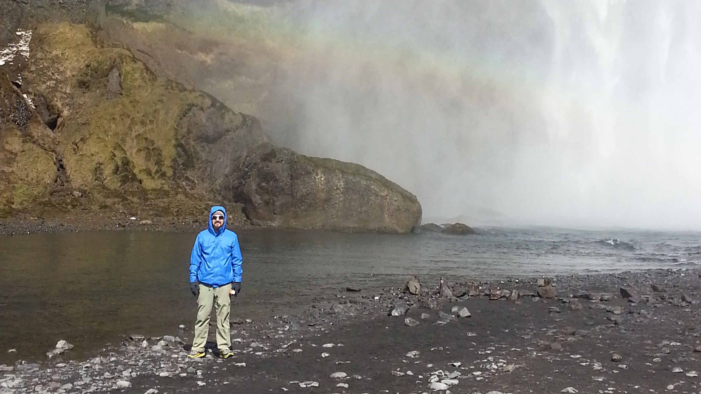 Clint stands in front of a waterfall with a rainbow forming behind him.