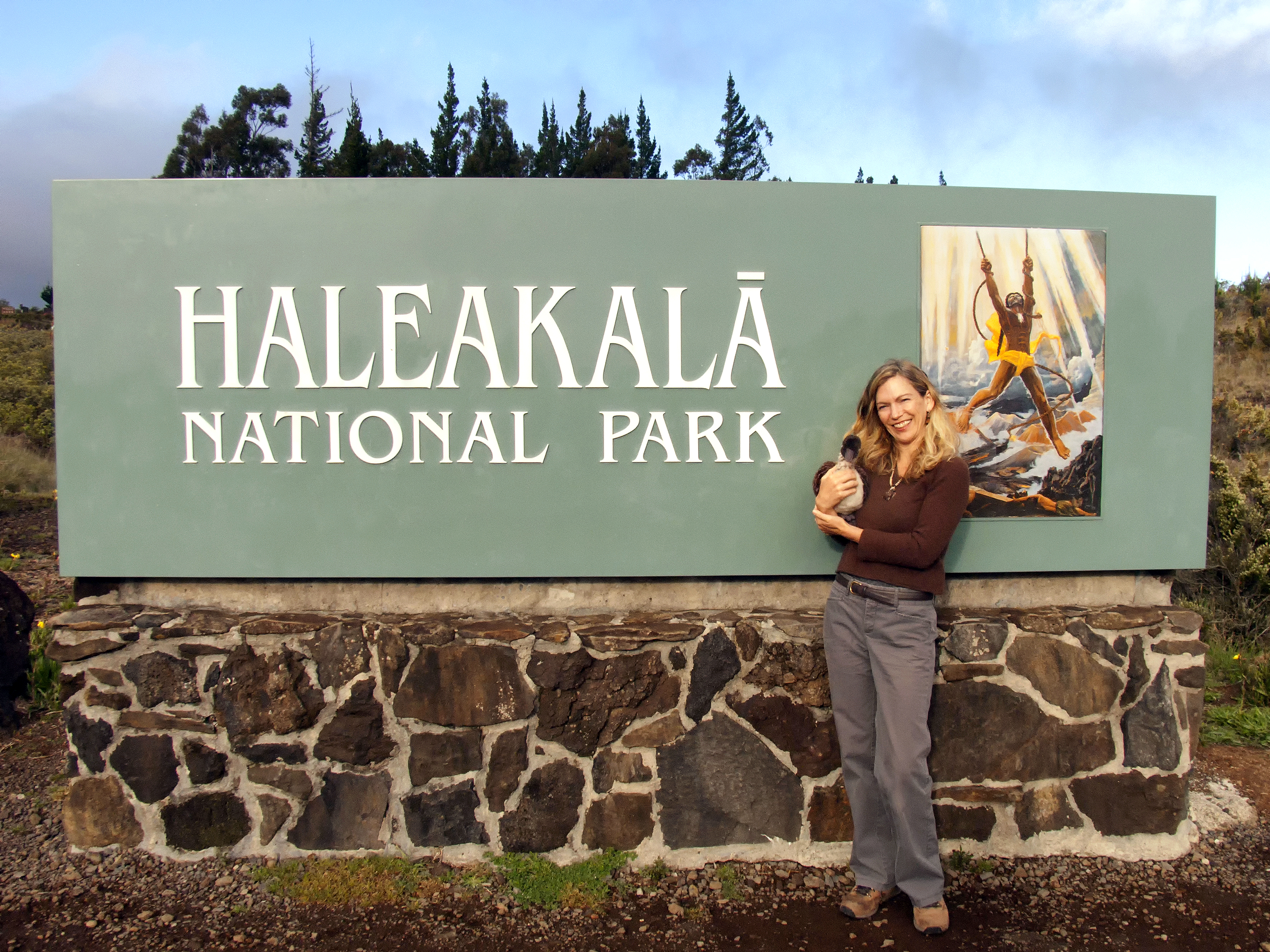 Wendy Hatch stands with Makana the plush nene in front of the Haleakala NP entry sign.